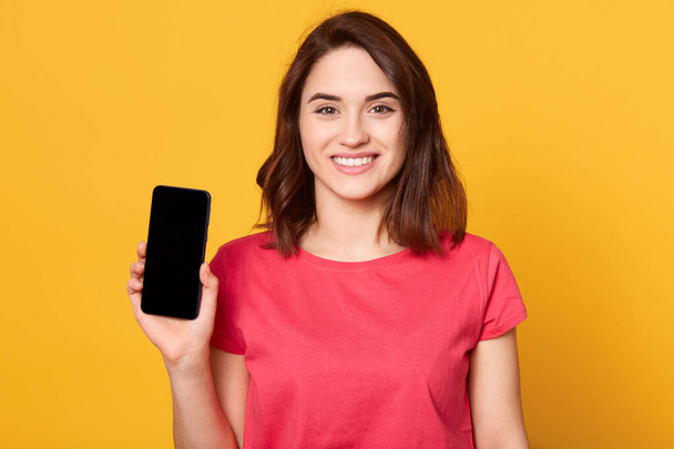 Horizontalshot of young woman showing her mobile cell phone with black screen over yellow background, adorable female wearing casual t shirt, looking smiling directlyt at camera. Copy space. - Фото, изображение