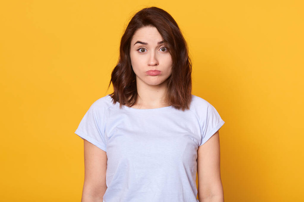 Studi oshot of dark haired young woman dresses casual white t shirt, standing and looking at camera, posing with pouty lips isolated over yellow background, being offended and having bad mood. - Photo, image