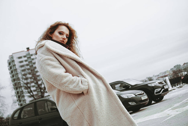 A girl with red curly hair in white coat poses on outdoor parking in cold autumn - Foto, Imagem