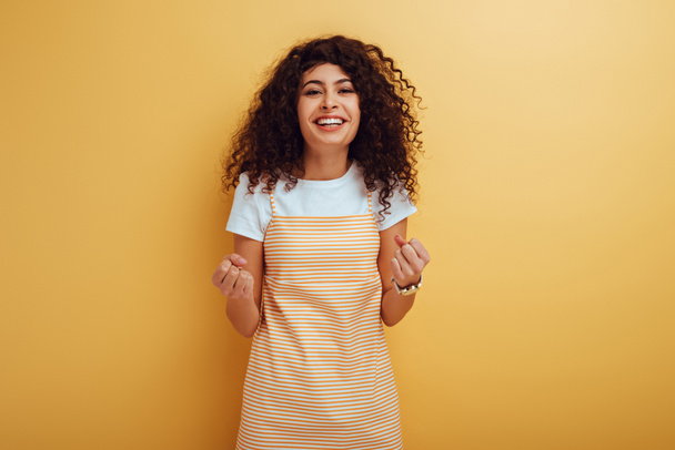 happy mixed race girl showing winner gesture while looking at camera on yellow background - Photo, Image