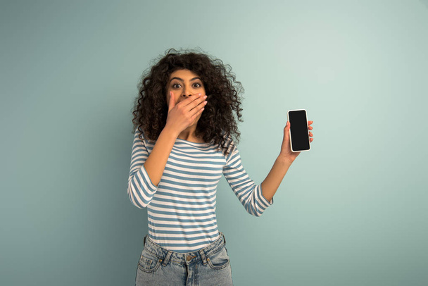 шокированная двурасовая девушка, прикрывающая рот рукой, показывая смартфон с пустым экраном на сером фоне
 - Фото, изображение
