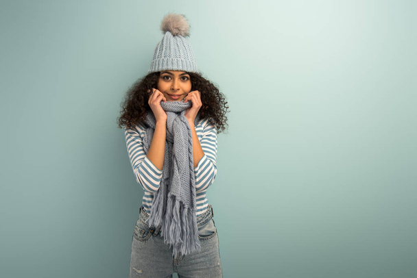hübsches zweirassiges Mädchen mit warmer Mütze und Schal, das vor grauem Hintergrund in die Kamera lächelt - Foto, Bild