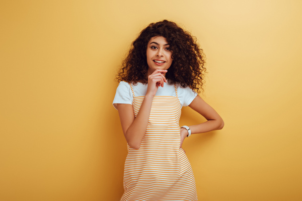 smiling bi-racial girl looking at camera while standing with hand on hip on yellow background - Photo, Image