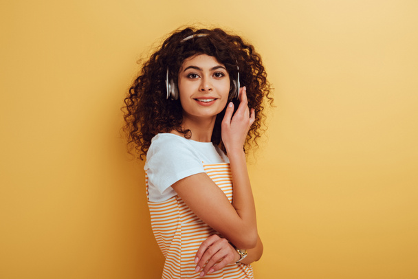 χαμογελαστό bi-φυλετικό κορίτσι σε ασύρματα ακουστικά κοιτάζοντας κάμερα σε κίτρινο φόντο - Φωτογραφία, εικόνα