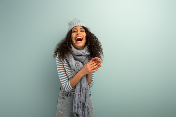 excitada chica bi-racial en sombrero caliente y bufanda riendo sobre fondo gris
 - Foto, Imagen