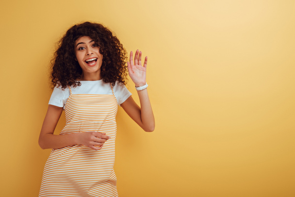 happy mixed-race girl waving hand while looking at camera on yellow background - Photo, Image