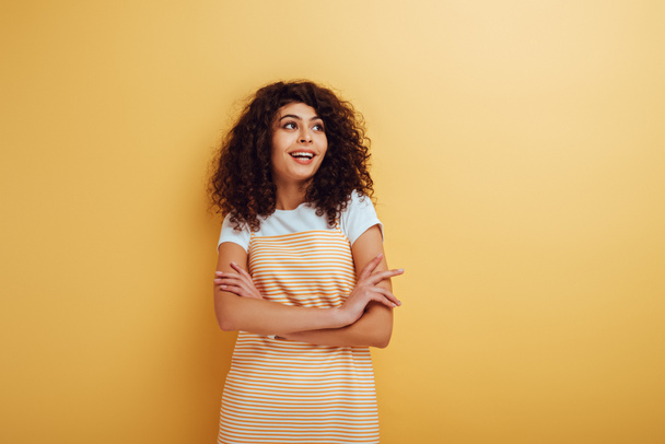 smiling, dreamy bi-racial girl standing with crossed arms and looking away on yellow background - Φωτογραφία, εικόνα