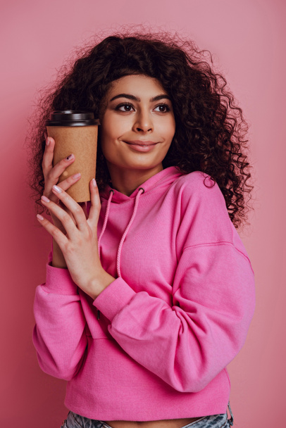 glimlachende, dromerige bi-raciale meisje houden koffie te gaan en weg te kijken op roze achtergrond - Foto, afbeelding