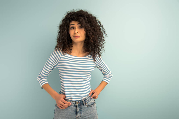 skeptical mixed girl looking at camera while holding hands on hips on grey background - Foto, Imagen