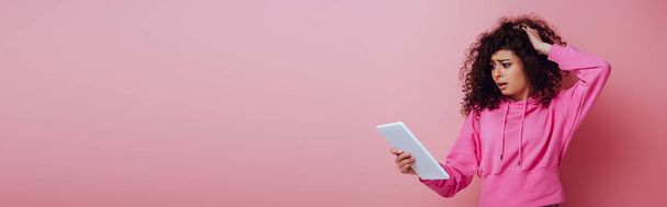 panoramic shot of disappointed bi-racial girl touching hair while looking at digital tablet on pink background - Photo, Image