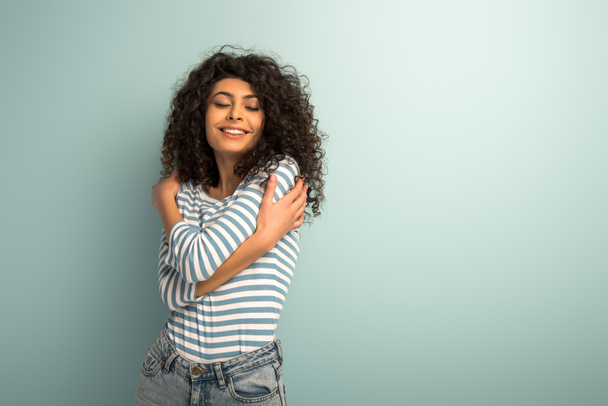 happy mixed race girl hugging herself while standing with closed eyes on grey background - Photo, Image