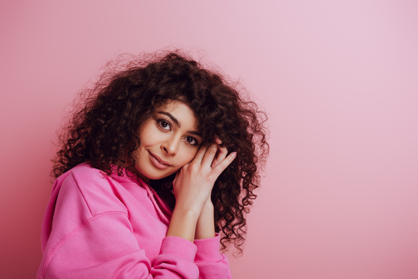 positive, dreamy bi-racial girl looking at camera while holding hands near face on pink background - Foto, Bild