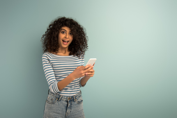 alegre bi-racial chica charlando en el teléfono inteligente mientras mira a la cámara en el fondo gris
 - Foto, Imagen