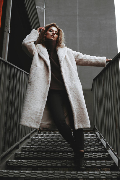 A girl with red curly hair in a white coat poses on the parking stairs - Fotoğraf, Görsel
