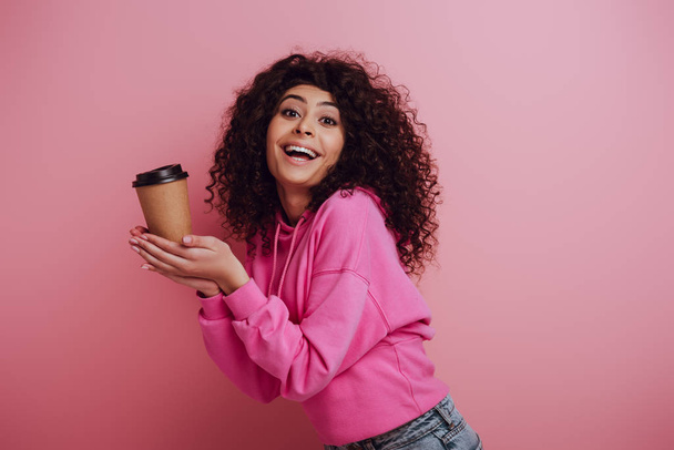 happy bi-racial girl smiling at camera while holding coffee to go on pink background - Fotoğraf, Görsel
