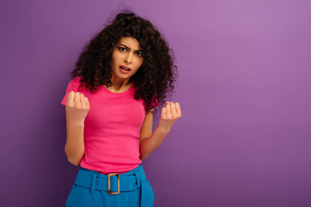 сердитая двурасовая девушка показывает кулаки, глядя в камеру на фиолетовом фоне
 - Фото, изображение
