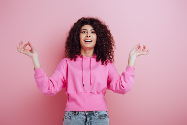 cheerful bi-racial girl looking at camera while standing in meditation pose on pink background - Fotografie, Obrázek