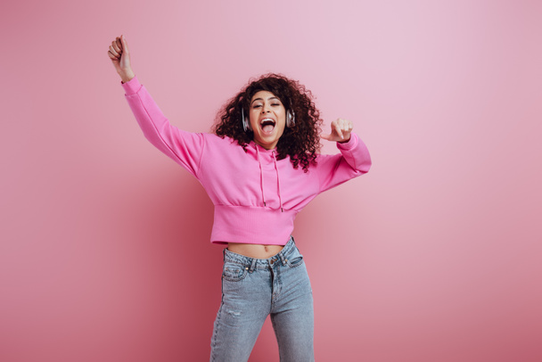 excited mixed race girl dancing and singing while listening music in wireless headphones on pink background - Photo, Image
