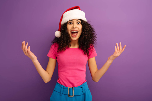 amazed bi-racial girl in santa hat looking at camera while standing with open arms on purple background - Foto, immagini