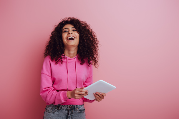 happy bi-racial woman laughing at camera while holding digital tablet on pink background - Фото, изображение