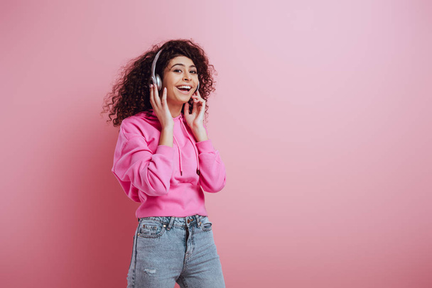 happy bi-racial girl smiling at camera while listening music in wireless headphones on pink background - Foto, Bild
