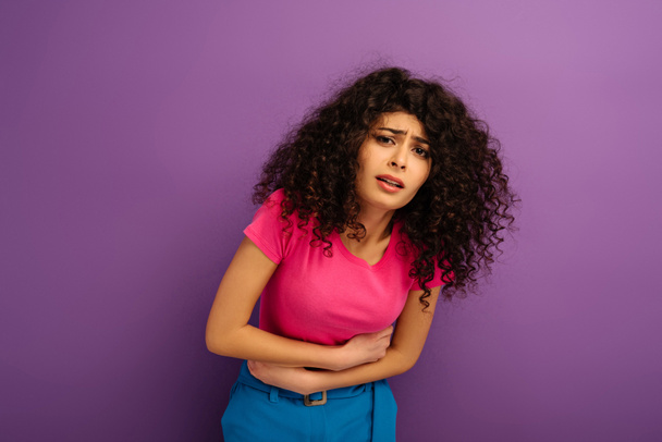 upset bi-racial girl touching stomach while suffering from abdominal pain on purple background - Photo, Image
