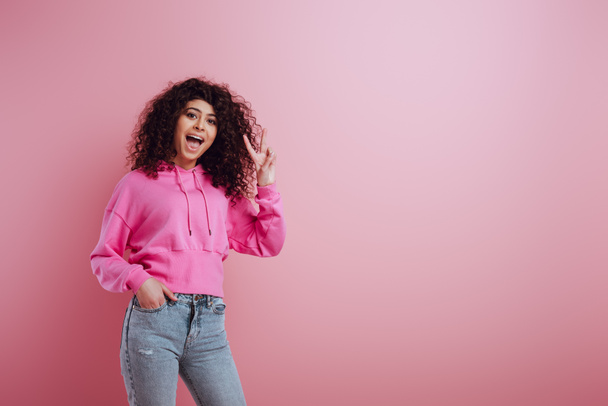 cheerful bi-racial girl showing victory gesture while holding hand in pocket on pink background - Fotó, kép