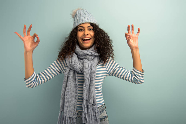 happy bi-racial girl in warm hat and scarf showing okay signs while looking at camera on grey background - Fotó, kép