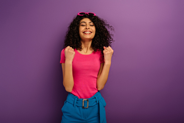 excitada chica bi-racial mostrando el gesto ganador mientras está de pie con los ojos cerrados sobre el fondo púrpura
 - Foto, Imagen