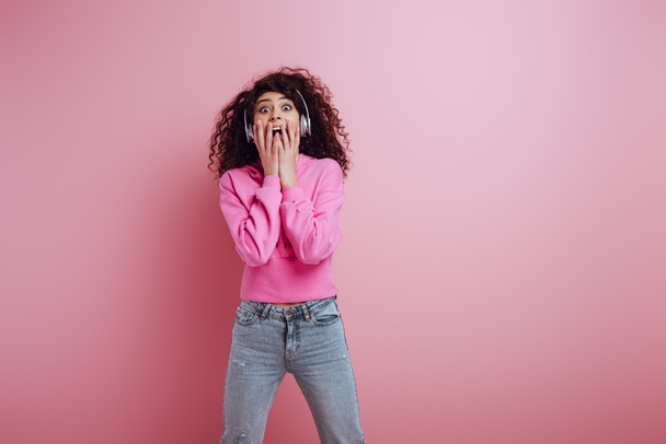 shocked bi-racial girl in wireless headphones covering open mouth with nads on pink background - Photo, Image