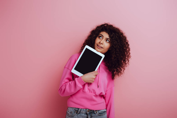 dreamy bi-racial girl looking away while showing digital tablet with blank screen on pink background - Foto, Bild