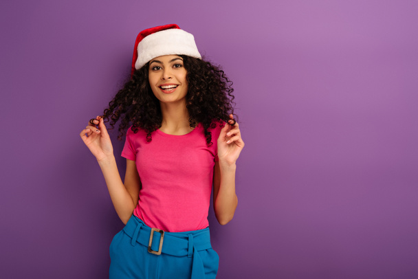 atractiva chica de raza mixta en sombrero de santa tocando el pelo mientras sonríe a la cámara sobre fondo púrpura
 - Foto, Imagen