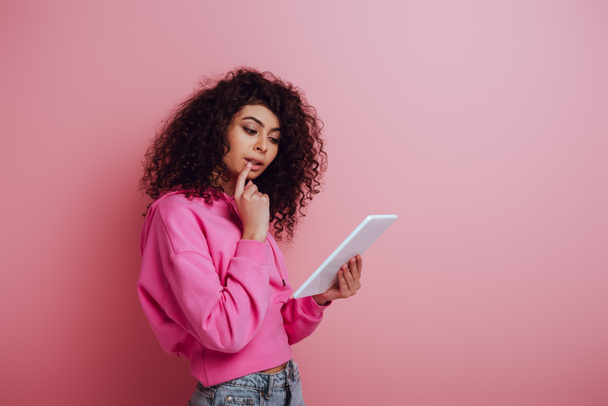 thoughtful bi-racial girl touching lip while using digital tablet on pink background - Fotoğraf, Görsel