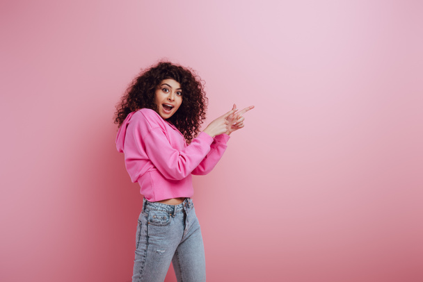 shocked bi-racial girl looking at camera while pointing with fingers on pink background - Zdjęcie, obraz