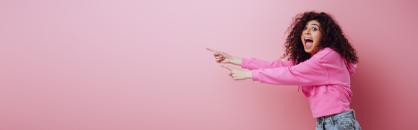 panoramic shot of shocked bi-racial girl looking at camera while pointing with fingers on pink background - 写真・画像
