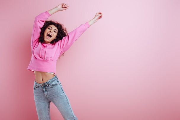 ευτυχισμένη bi-φυλετικό κορίτσι κοιτάζοντας κάμερα, ενώ στέκεται με υψωμένα τα χέρια σε ροζ φόντο - Φωτογραφία, εικόνα