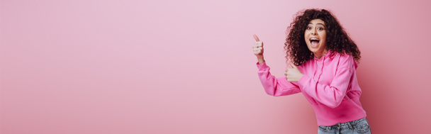πανοραμική φωτογραφία του ενθουσιασμένοι bi-φυλετικό κορίτσι δείχνει αντίχειρες επάνω σε ροζ φόντο - Φωτογραφία, εικόνα