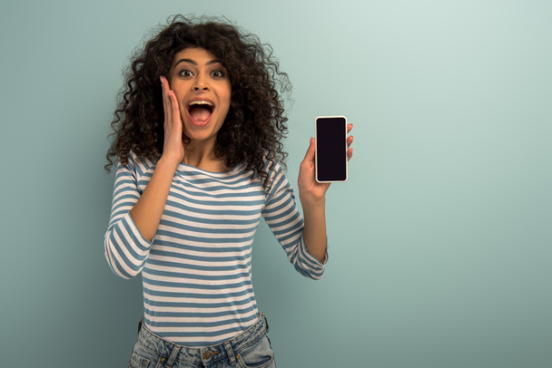 excitée fille bi-raciale regardant la caméra tout en montrant smartphone avec écran vide sur fond gris
 - Photo, image