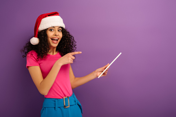 cheerful mixed race girl in santa hat pointing with finger at digital tablet on purple background - Foto, immagini