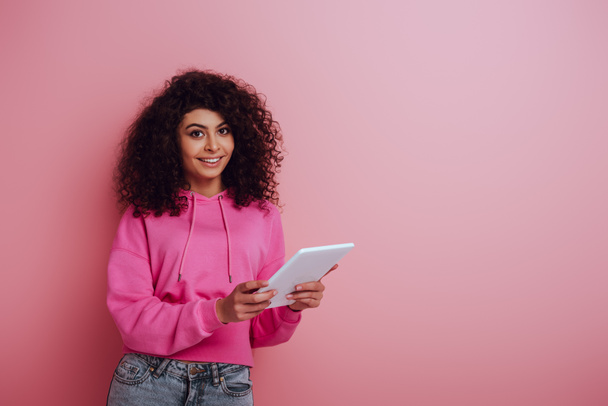 cheerful mixed race girl smiling at camera while using digital tablet on pink background - Foto, immagini