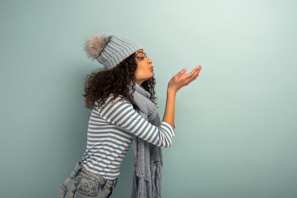  vista lateral de chica bi-racial en sombrero caliente y bufanda enviando beso de aire sobre fondo gris
 - Foto, imagen