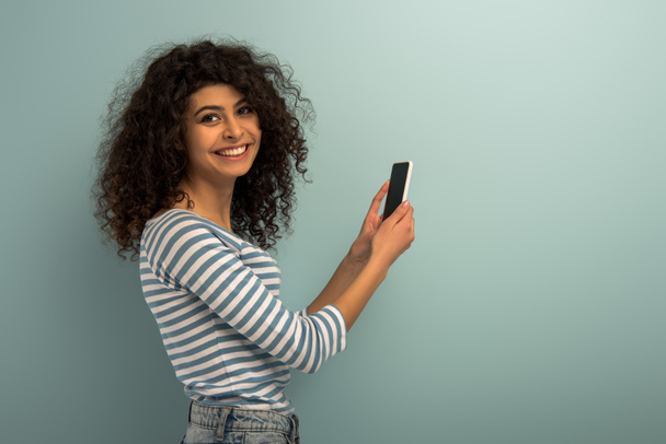 boldog bi-rasszista lány mosolyog a kamera, miközben gazdaság smartphone üres képernyő szürke háttér - Fotó, kép