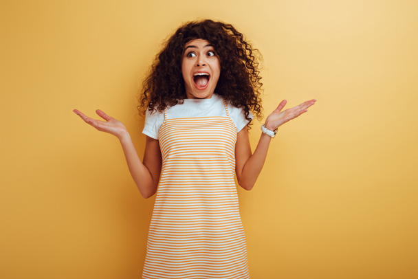 shocked mixed race girl looking away while standing with open arms on yellow background - Photo, Image