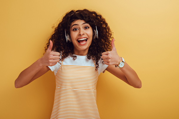 excited mixed race girl in wireless headphones showing thumbs up on yellow background - Foto, Bild