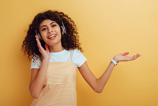 cheerful mixed race girl in wireless headphones smiling at camera on yellow background - Foto, afbeelding