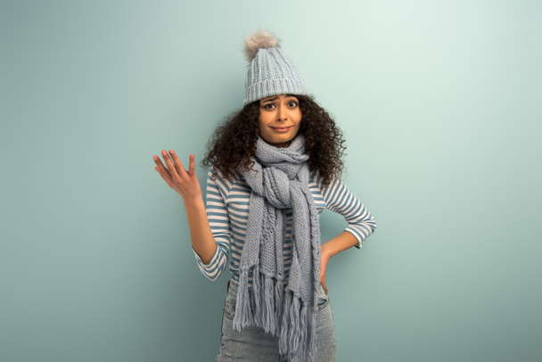 beledigd bi-raciale meisje in warme hoed en sjaal kijken naar camera op grijze achtergrond - Foto, afbeelding