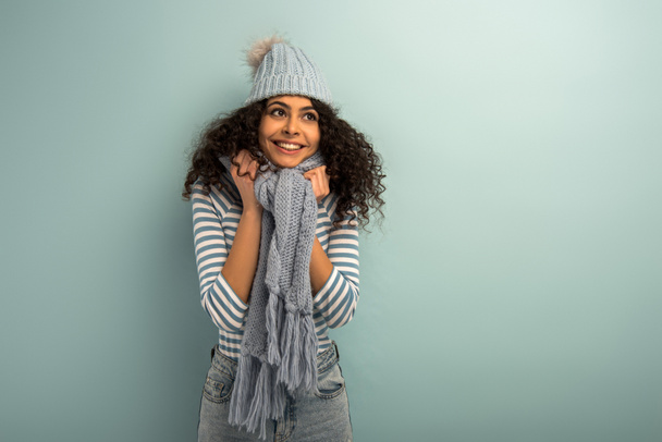 bonita chica bi-racial en sombrero caliente y bufanda sonriendo y mirando hacia otro lado sobre fondo gris
 - Foto, imagen