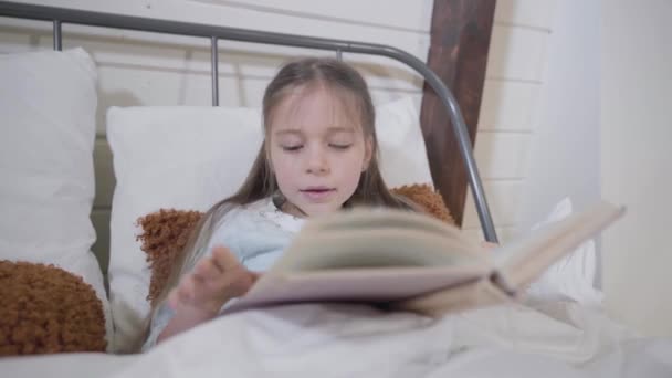 Portrait of pretty Caucasian girl lying in bed and reading book. Smart child going for sleep in the evening. Bedtime, hobby, leisure. - Video
