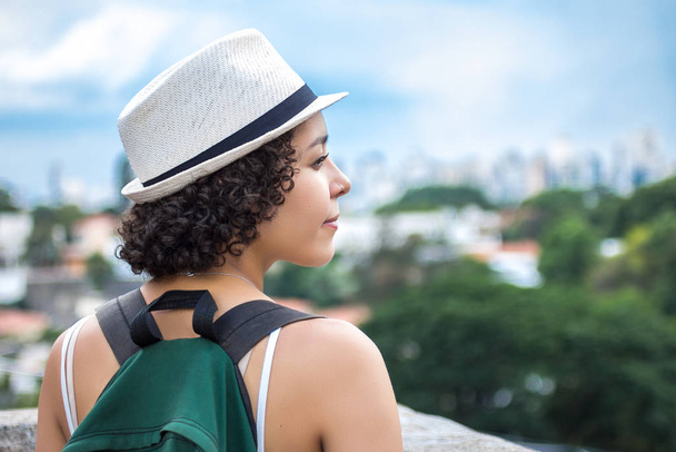 Jonge reiziger op panoramisch uitzicht op stedelijk gebied. Draag witte hoed en groene rugzak. - Foto, afbeelding