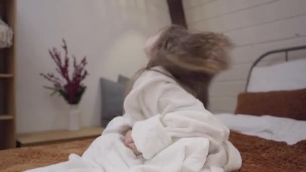 Camera approaching to cute little Caucasian girl in white bathrobe shaking long curly hair. Pretty kid sitting on bed after shower and smiling. Leisure, lifestyle, relaxation. - Metraje, vídeo
